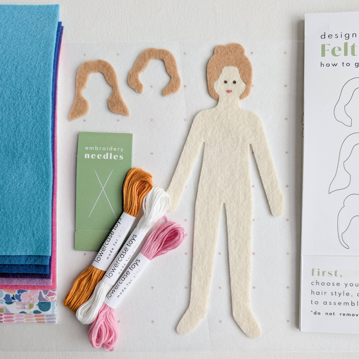 Felt craft kit with a paper doll, felt pieces, embroidery threads, and a book on a white surface.