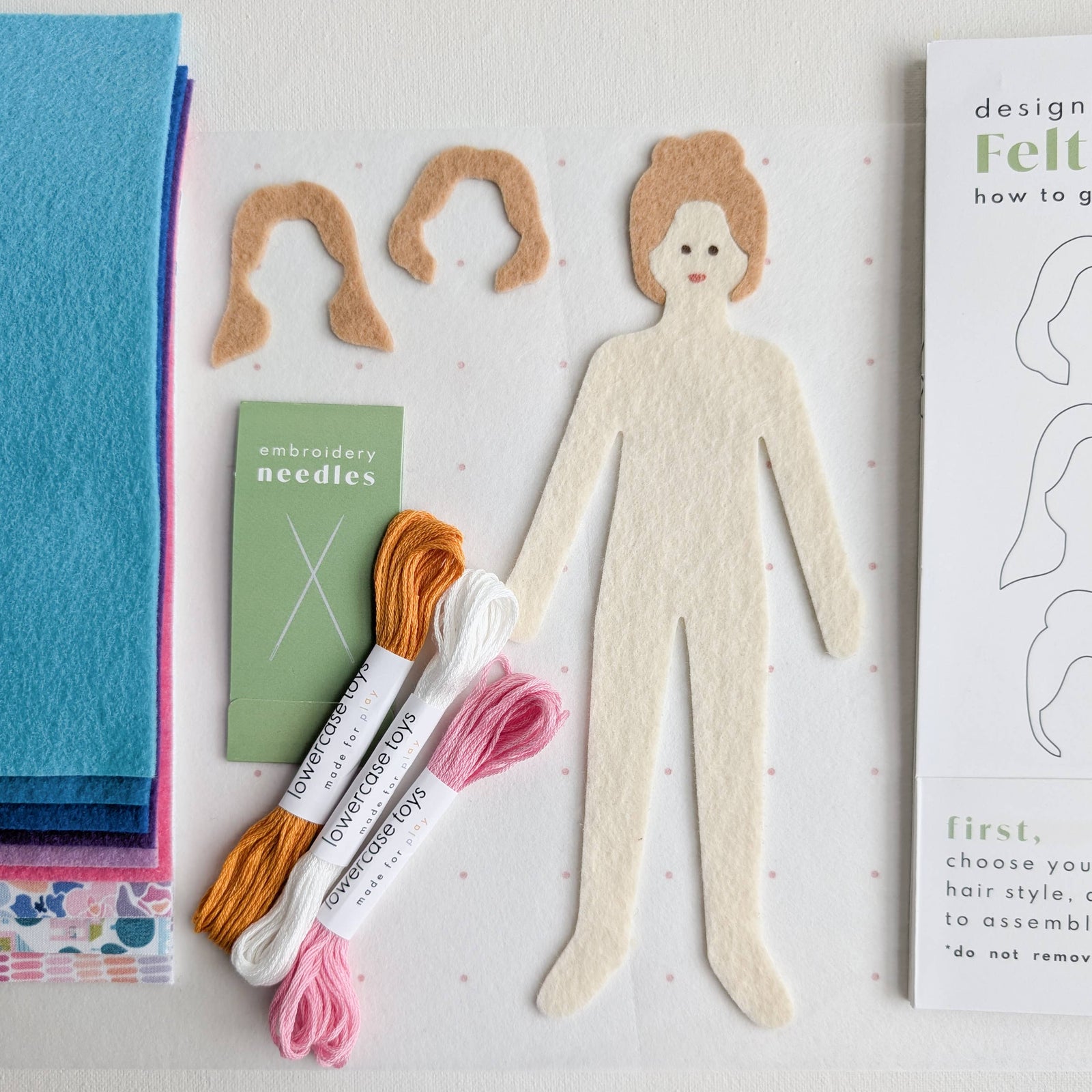 Felt doll design set with packaging, felt pieces, needles, and a paper doll template on a white background.