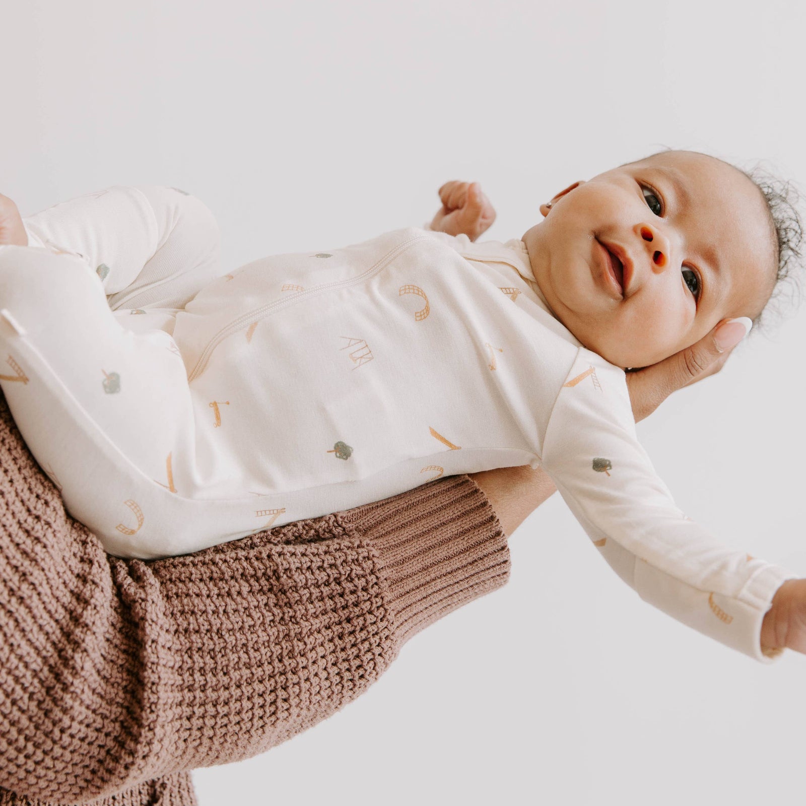 Baby in a beige onesie with play ground prints sitting on a textured blanket against a light blue background