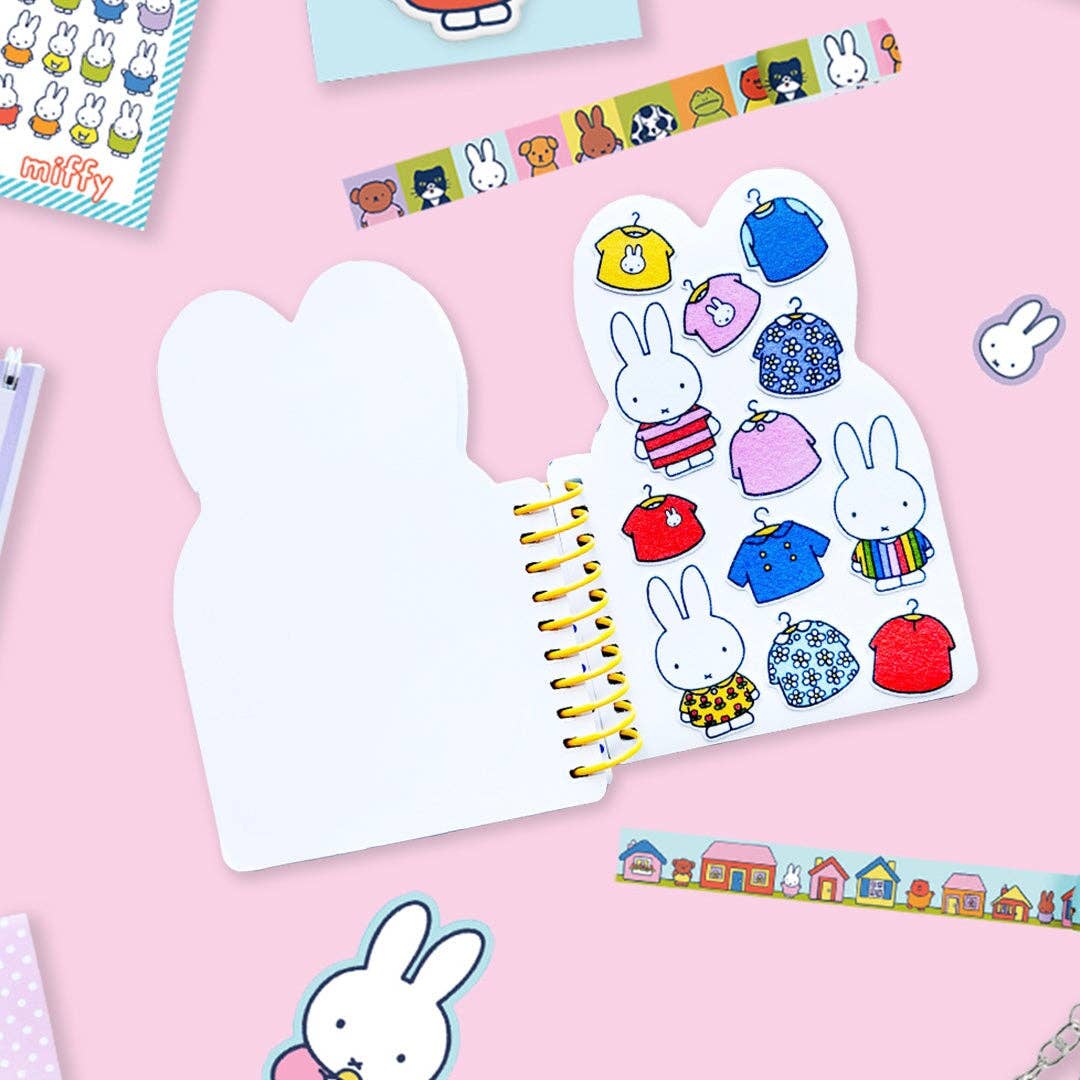 A white spiral-bound notebook with bunny-shaped covers, shown open on a pink background. The right cover features cartoon rabbit characters wearing colorful clothing, and the left cover is blank.