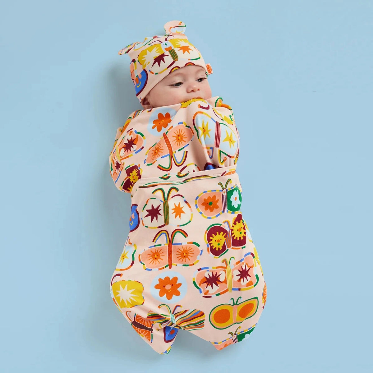A baby wrapped in a colorful floral and butterfly-patterned swaddle blanket, wearing a matching hat, against a solid light blue background.