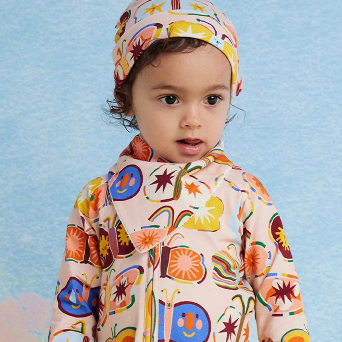 A toddler wearing a matching patterned outfit with and a matching bib and hat featuring a colorful butterfly print, set against a textured light blue background.