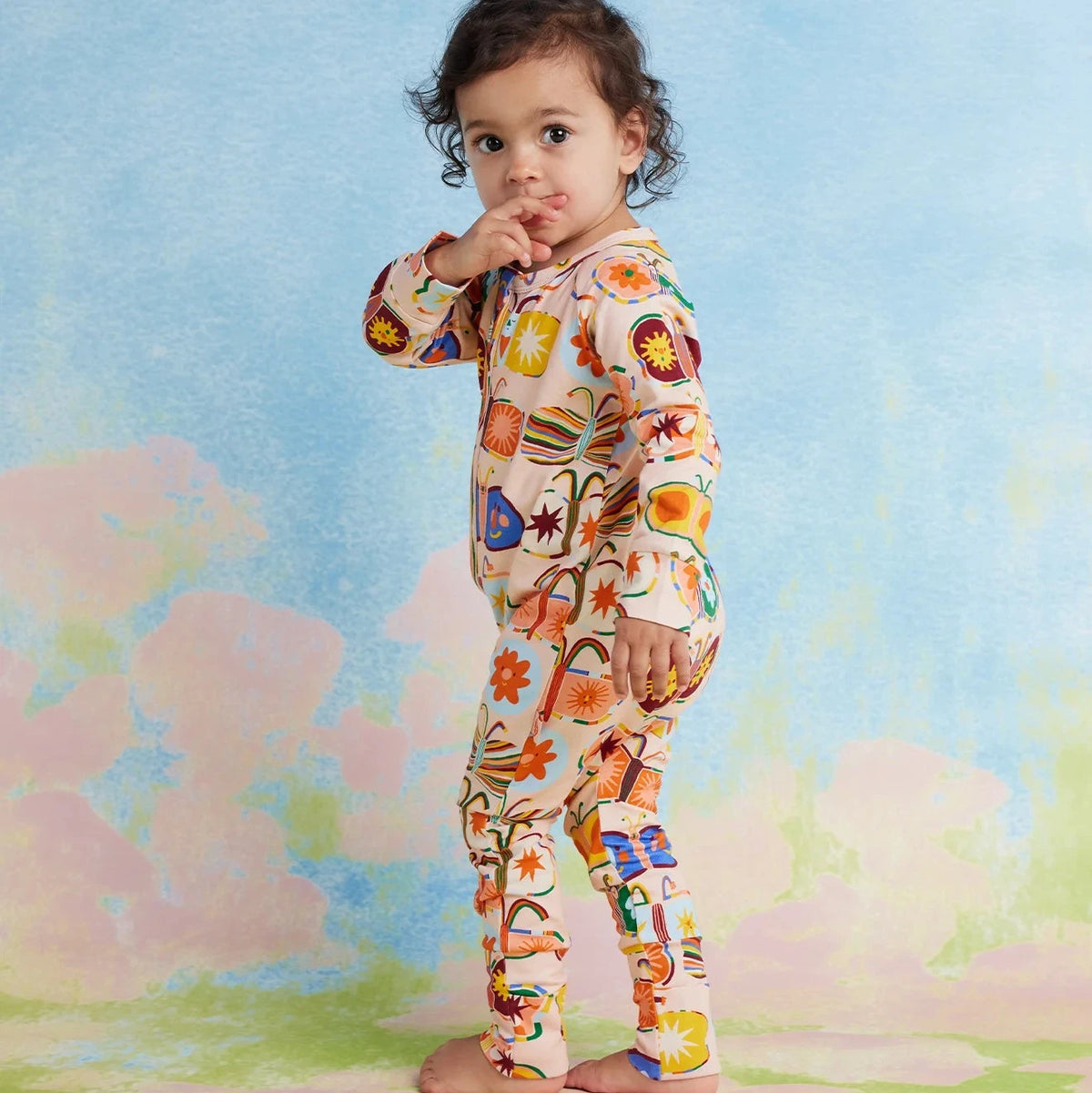 A toddler stands barefoot in a studio setting, wearing a one-piece romper with butterfly pattern on a cream base. The background is a soft pastel blue, pink, and green painted backdrop.