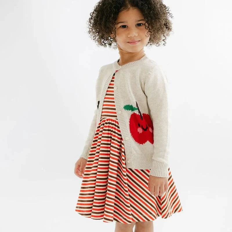 A young child stands barefoot against a plain white studio background, wearing a cream knit cardigan over a red and white striped dress with a red apple applique on the front.