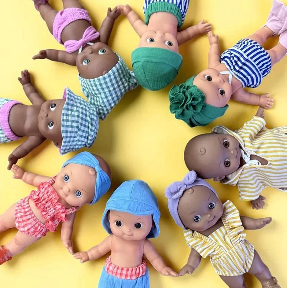 Baby dolls with skin tones are arranged in a circle on a bright yellow background, wearing colorful striped and gingham outfits with matching hats and bows.