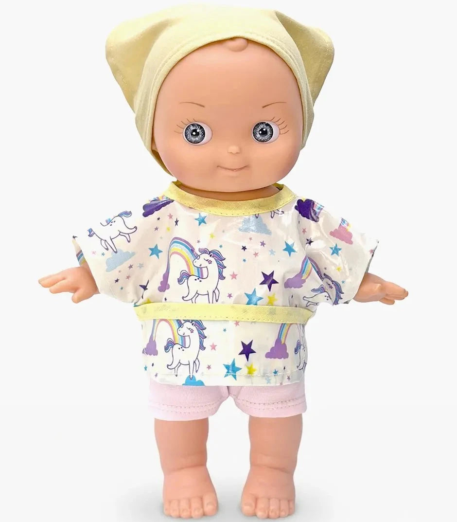 A doll with blue eyes wearing a pale yellow bonnet and a short-sleeve top and shorts set with a unicorn and rainbow print on a white background.