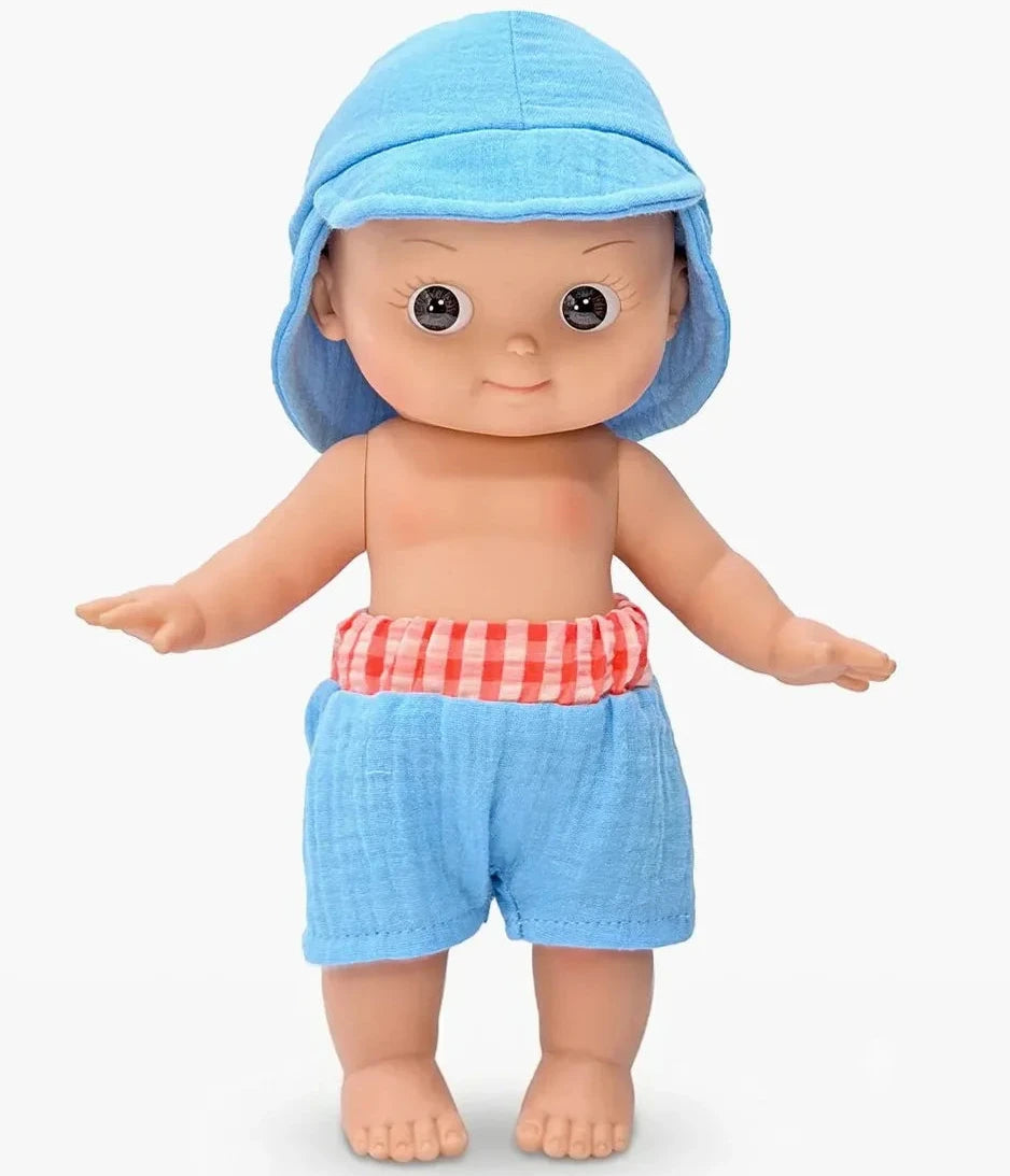A doll with large dark eyes, wearing a light blue sun hat and matching light blue shorts with a red-and-white gingham waistband, standing against a plain light gray background.