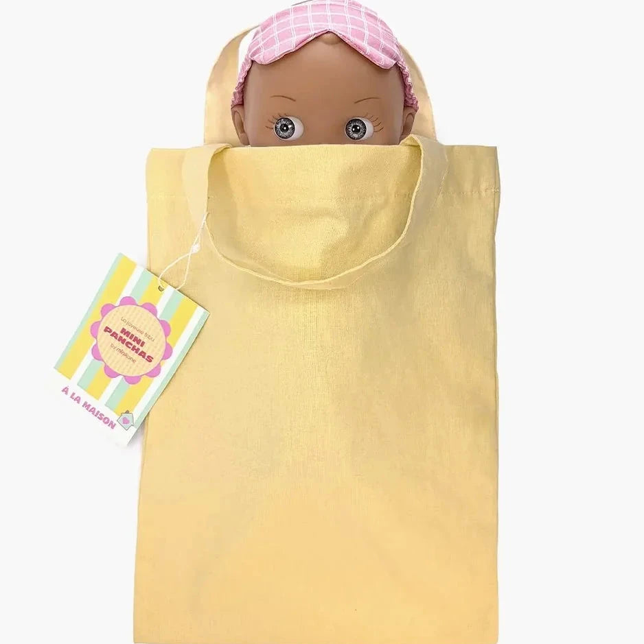 A pale yellow fabric bag shown with a doll peeking out wearing a pink checkered sleep cap. A colorful product tag is attached on the left side, set against a plain white background.