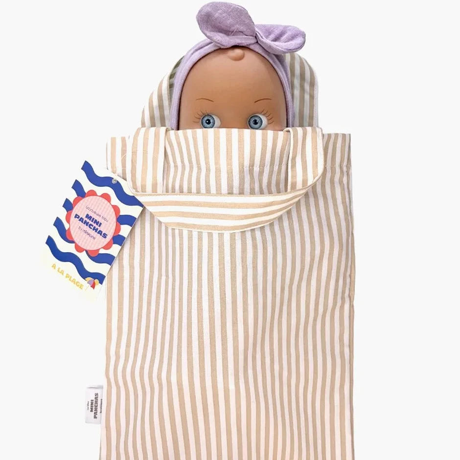 A baby doll peeking out from a cream and beige vertically striped fabric bag, with a lavender hair wrap on its head. A blue, white, and pink tag is attached to the left side of the wrap.