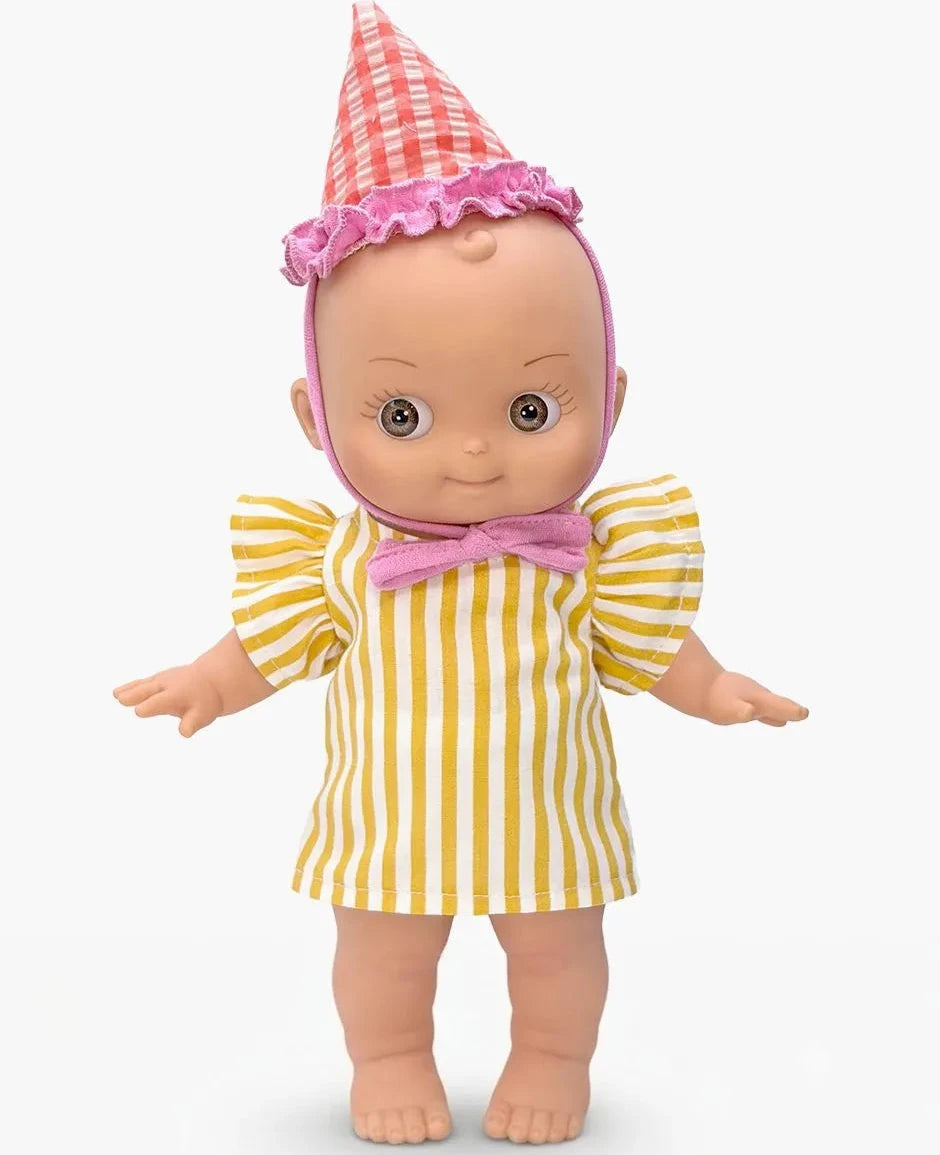 A baby doll with a pink and white checkered party hat, wearing a yellow and white striped dress with a pink bow tie, standing against a light gray background.