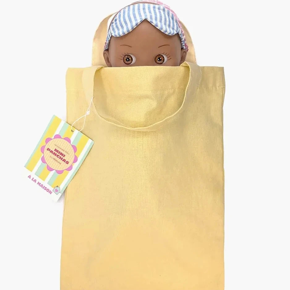 A pale yellow fabric  bag with a matching drawstring tie, featuring a hanging tag and a doll peeking out from the top wearing a blue and white striped sleep mask  set against a plain white background.