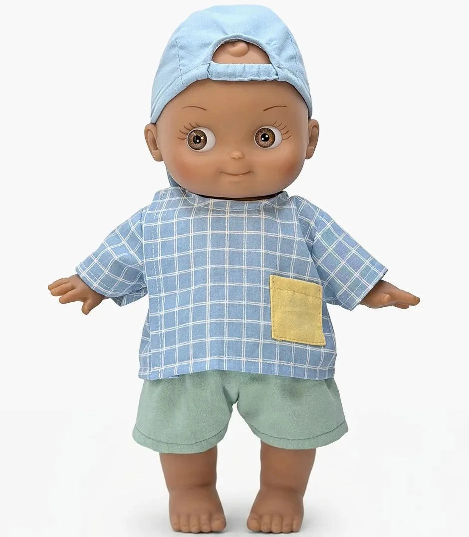 A doll with brown skin and large eyes wearing a light blue plaid shirt with a yellow pocket, a matching light blue cap, and light green shorts, standing against a plain white background.