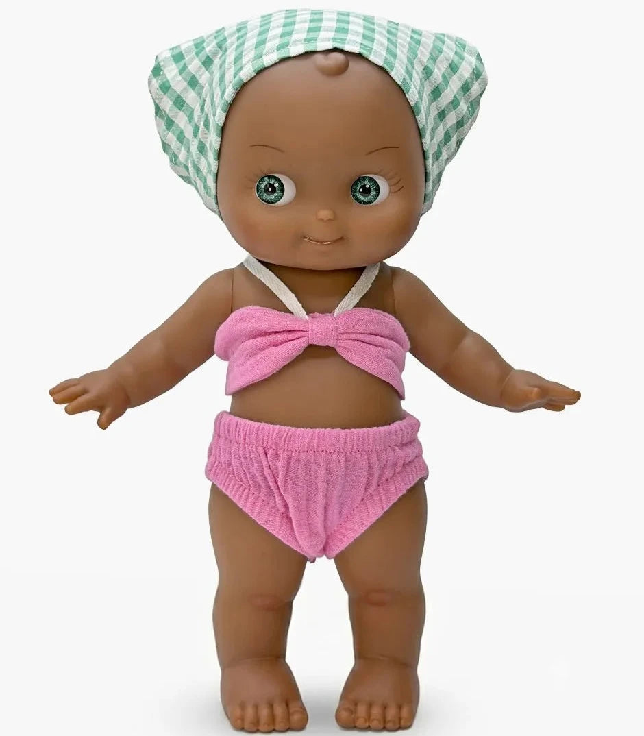 A baby doll with brown skin and large blue eyes, wearing a pink two-piece swimsuit with a bow top and matching bottoms, and a green and white checkered towel wrap on its head, set against a plain light gray background.