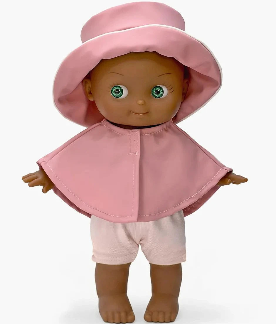 A doll with dark brown skin and large green eyes, wearing a pink wide-brim hat, a pink cape-style top, and light pink shorts, standing against a plain light gray background.