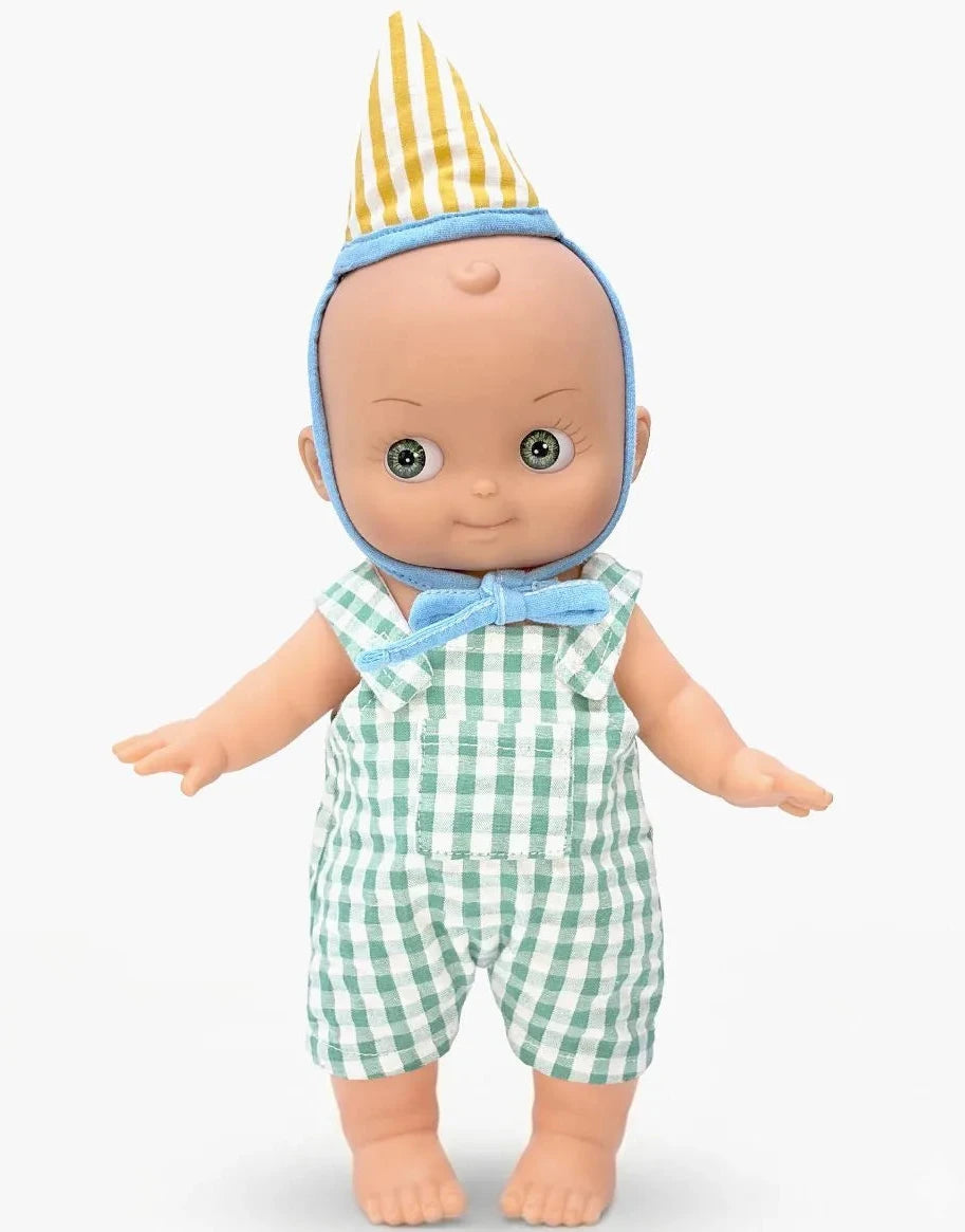 A baby doll with a bald head, large eyes, and a yellow-and-white striped party hat, wearing a green-and-white gingham romper with a light blue bow tie and blue chin strap, standing against a light gray background.
