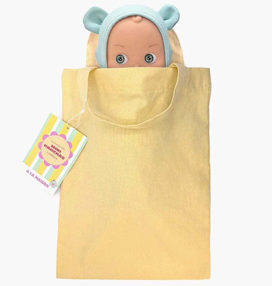 A pale yellow fabric gift bag with a drawstring handle, featuring a hanging tag and a peeking doll with a light blue bear-ear bonnet on a white background.