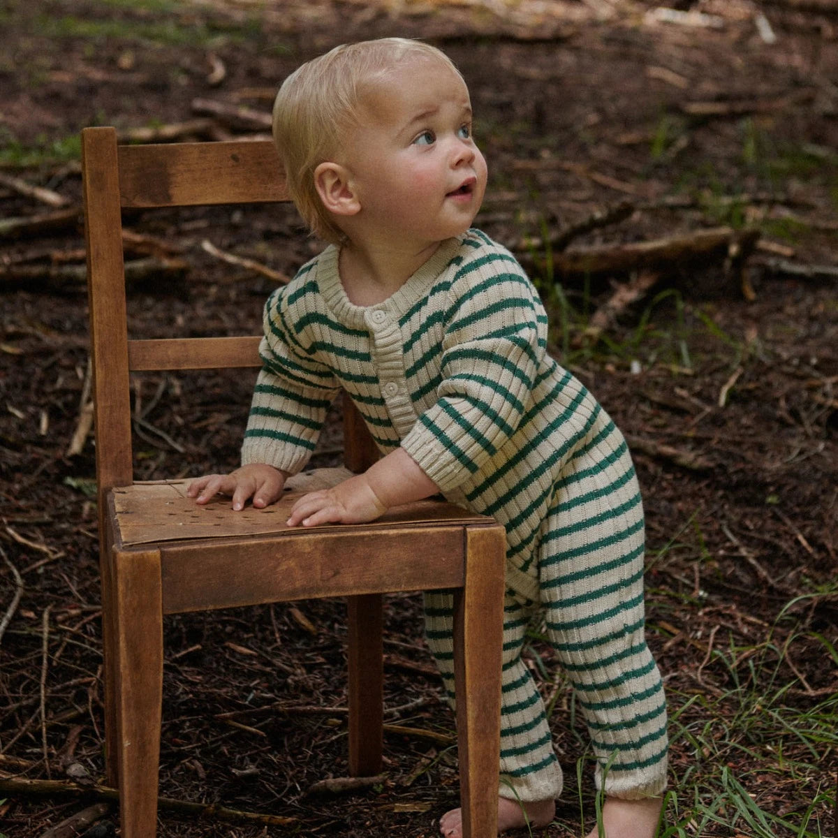 Child in a striped outfit standing next to a wooden chair outdoors.