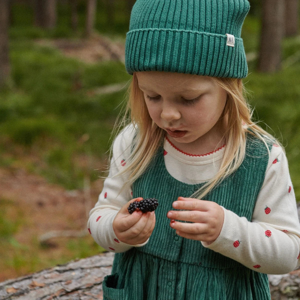 Child in a green hat holding a blackberry in a forest setting