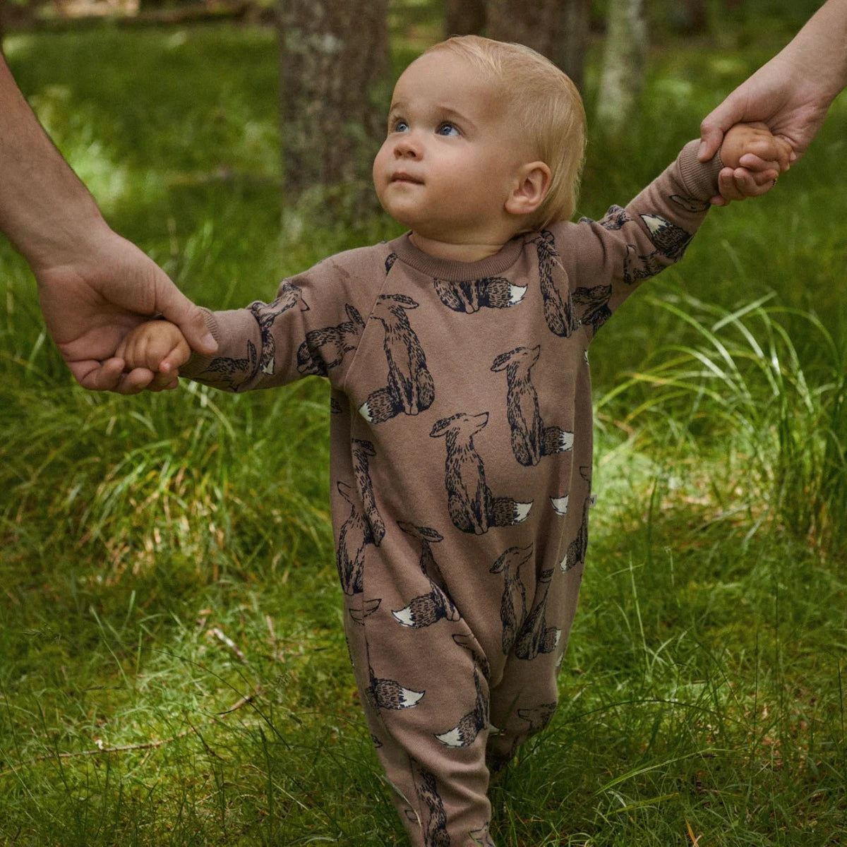 Baby in a brown onesie with Fox prints standing in a grassy area, holding hands with two adults.