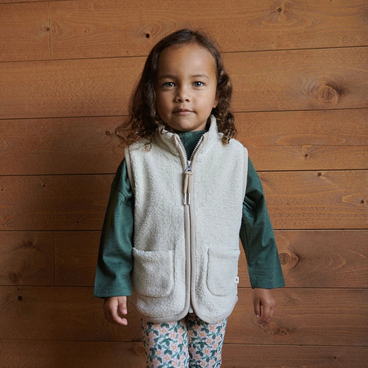 Child wearing an oatmeal vest over a green shirt against a wooden background