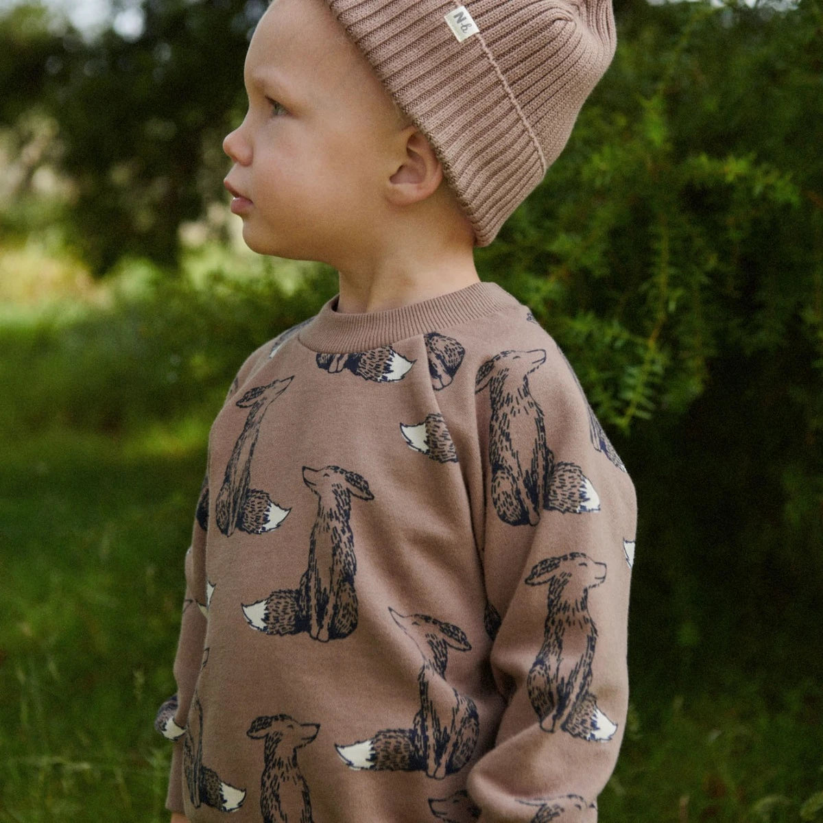 Child wearing a brown sweatshirt with fox patterns and a matching beanie outdoors.