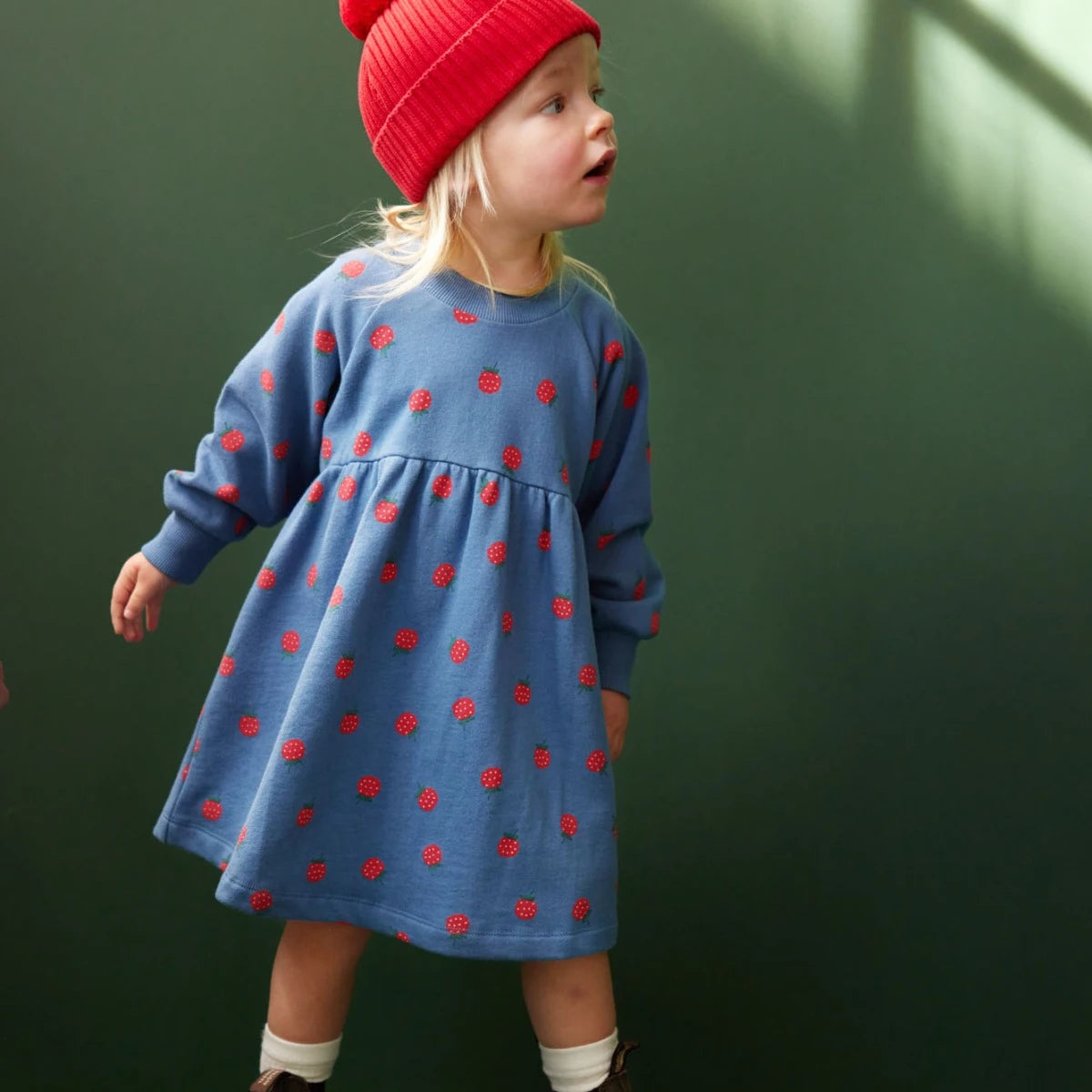 Child wearing a blue dress with red berries and a red beanie against a green background