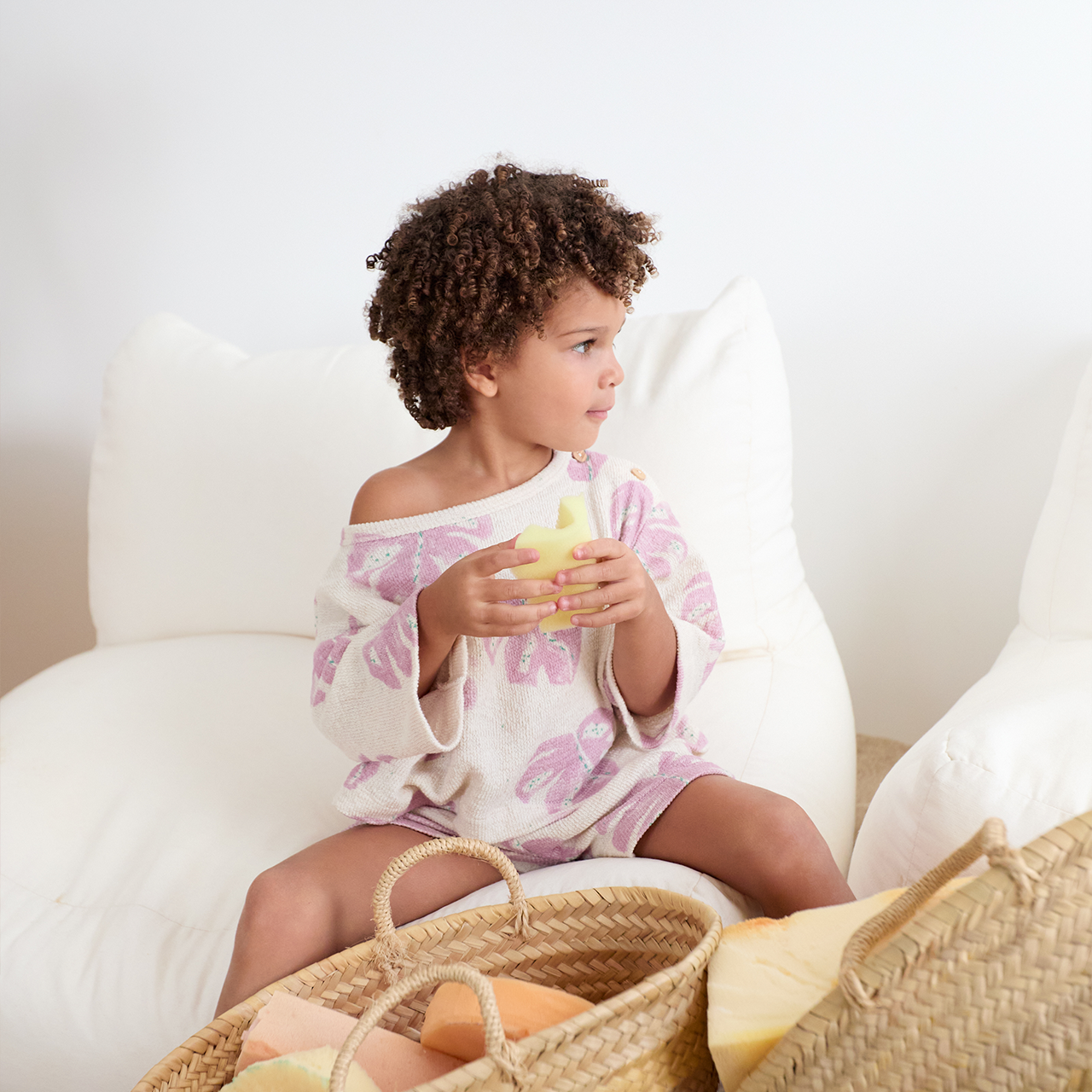 Child sitting on a white chair holding a yellow object with baskets nearby.