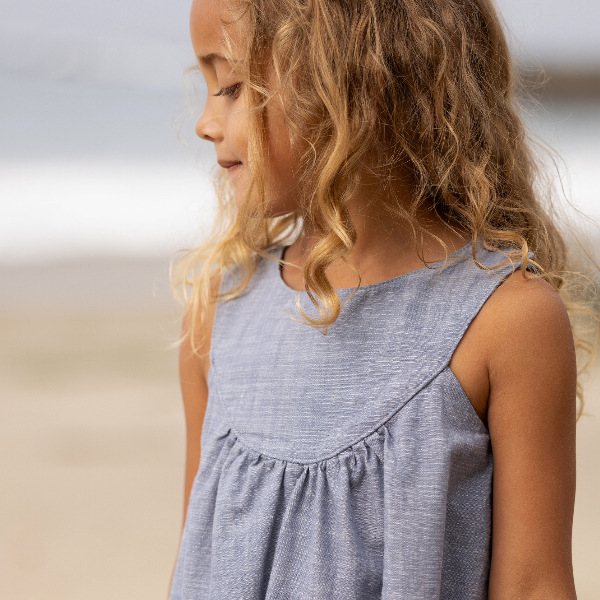 Young girl in a blue dress standing on a sandy beach with a large rock in the background.