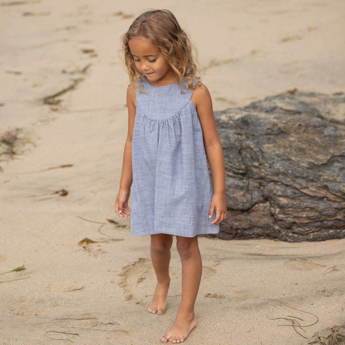 Young girl in a blue dress standing on a sandy beach with a large rock in the background.