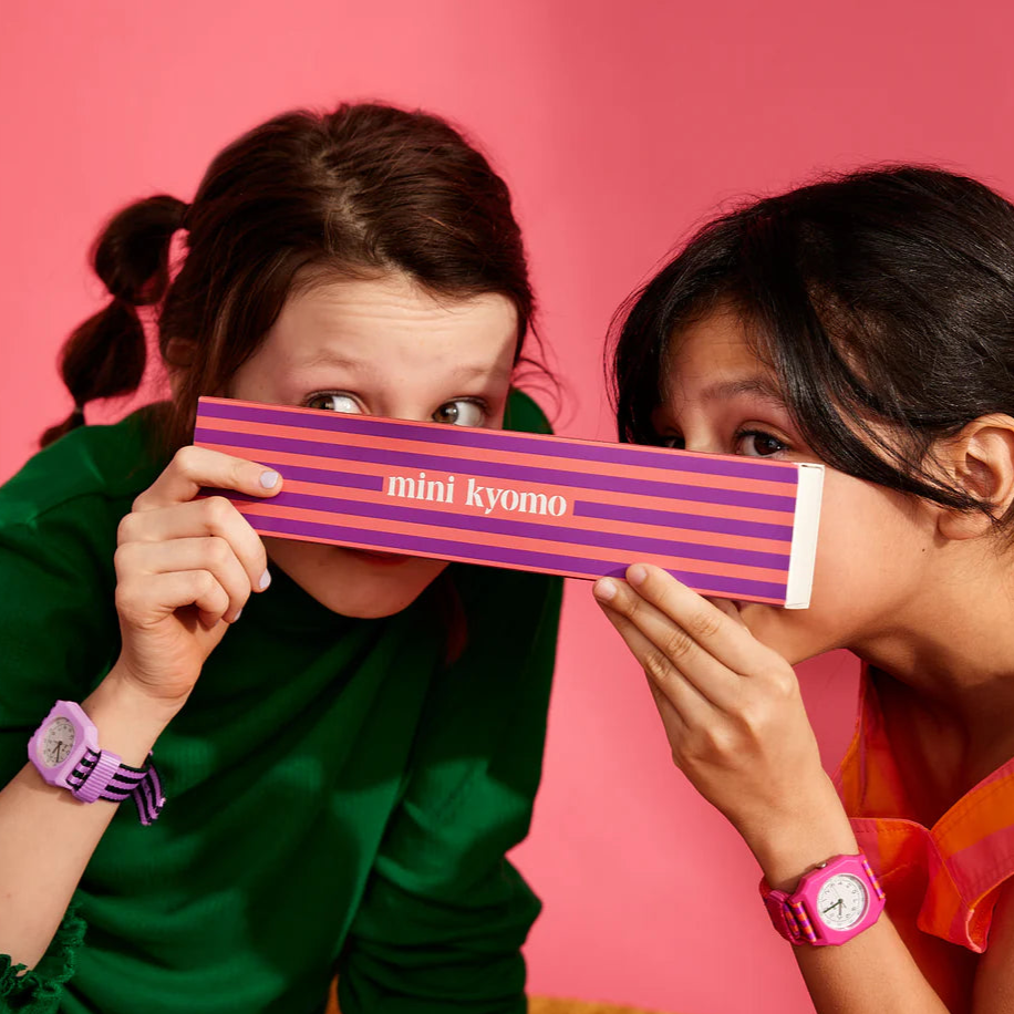 Two individuals holding a Mini Kyomo watch with a striped pattern, predominantly pink and purple, against a pink background.