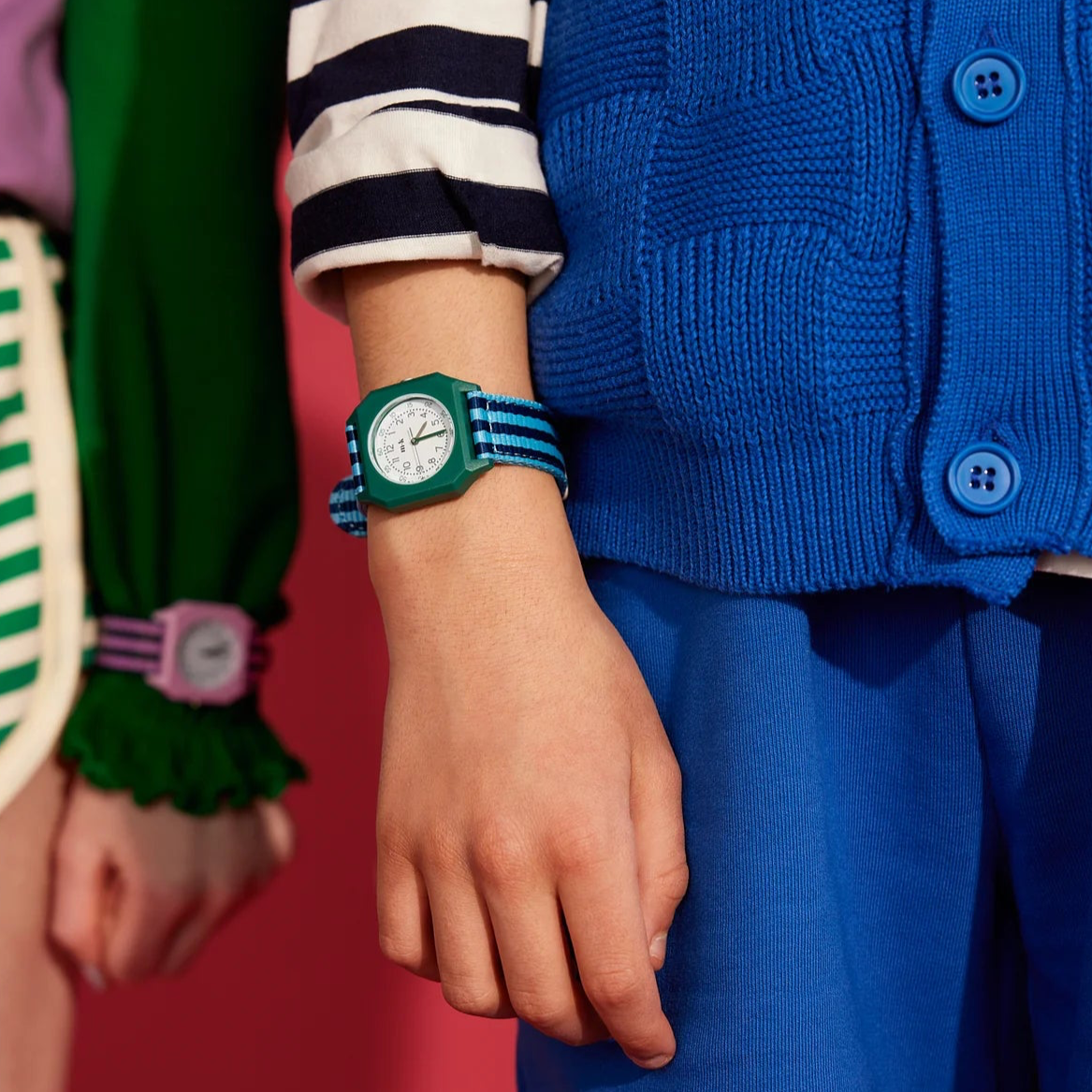 Two individuals holding a Mini Kyomo watch with a striped pattern, predominantly pink and purple, against a pink background.
