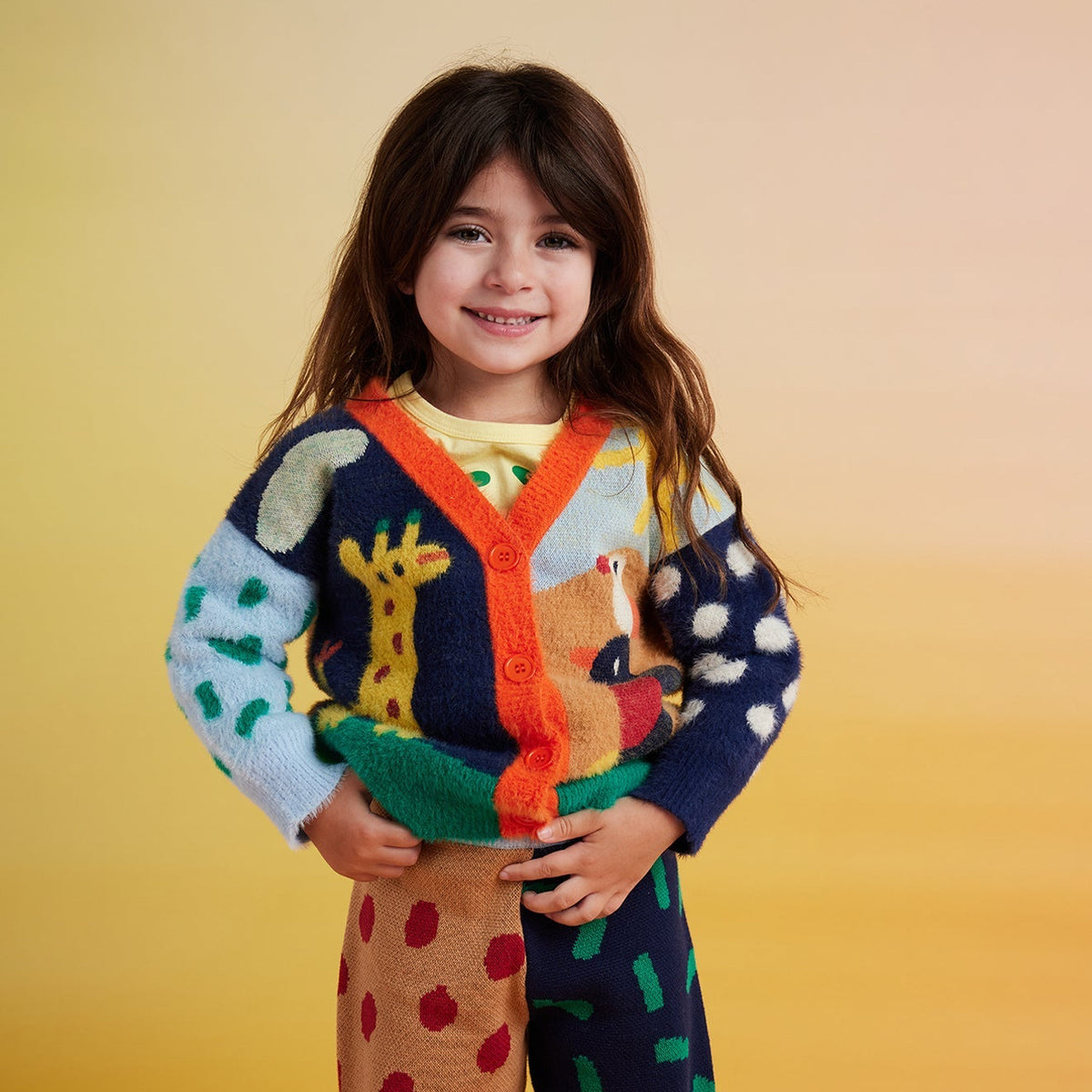 A young girl wearing a colorful knit cardigan and matching patterned pants against a warm yellow-to-orange gradient background.