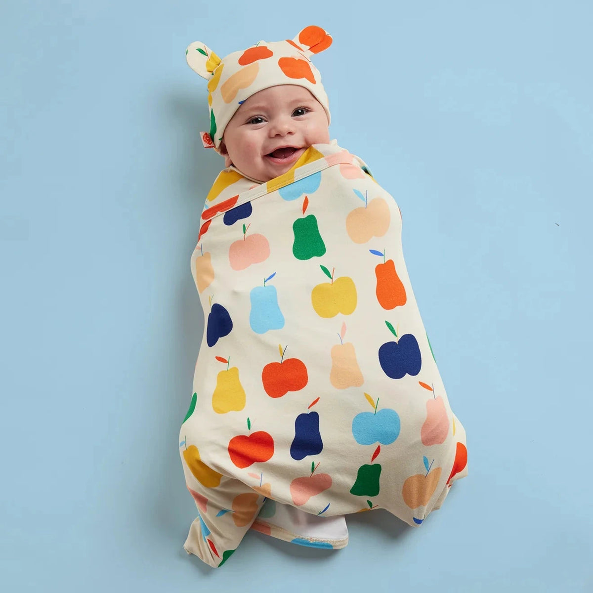 A baby wrapped in a cream-colored swaddle blanket with a colorful fruit print, wearing a matching hat, set against a solid light blue background.