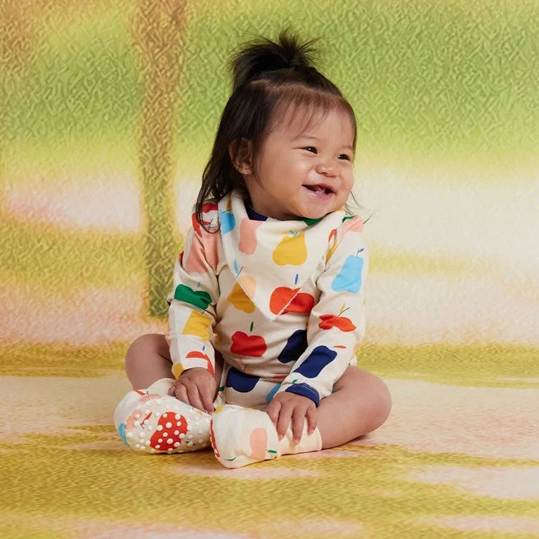 A baby sitting on a textured yellow and green backdrop, wearing a cream-colored long-sleeve bodysuit with a colorful pattern of red, blue, yellow, and green fruits  along with matching patterned socks.