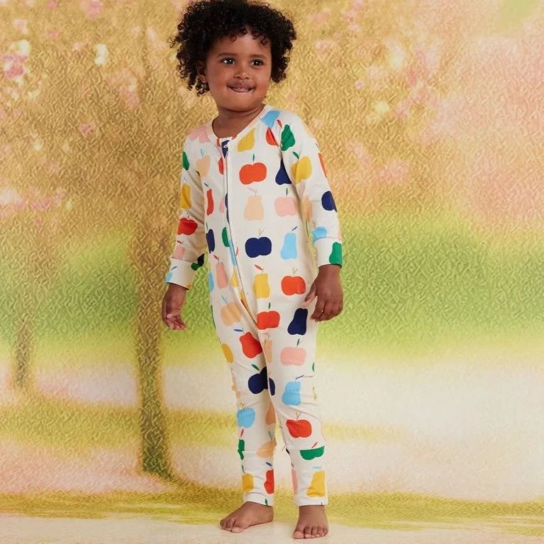 A toddler stands barefoot wearing a one-piece zip-up pajama top and pants with a colorful fruit print on a white base. The background is a soft, textured, warm-toned studio backdrop resembling a tree with pink and yellow leaves.