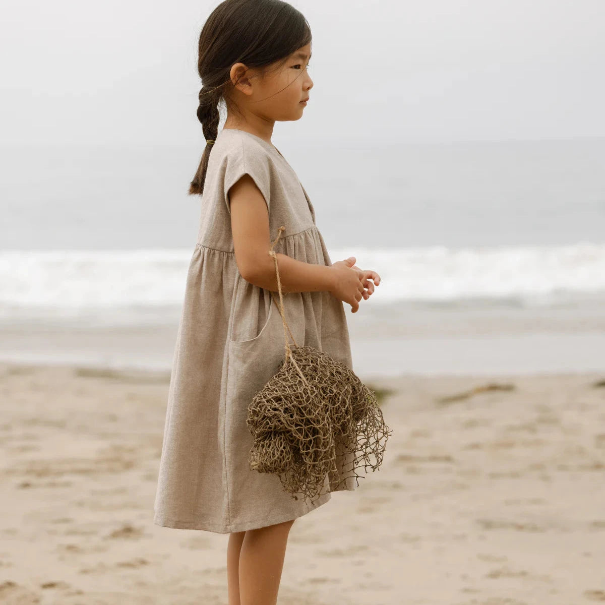 Young girl in a beige dress standing on a beach holding a bundle of dried grass.