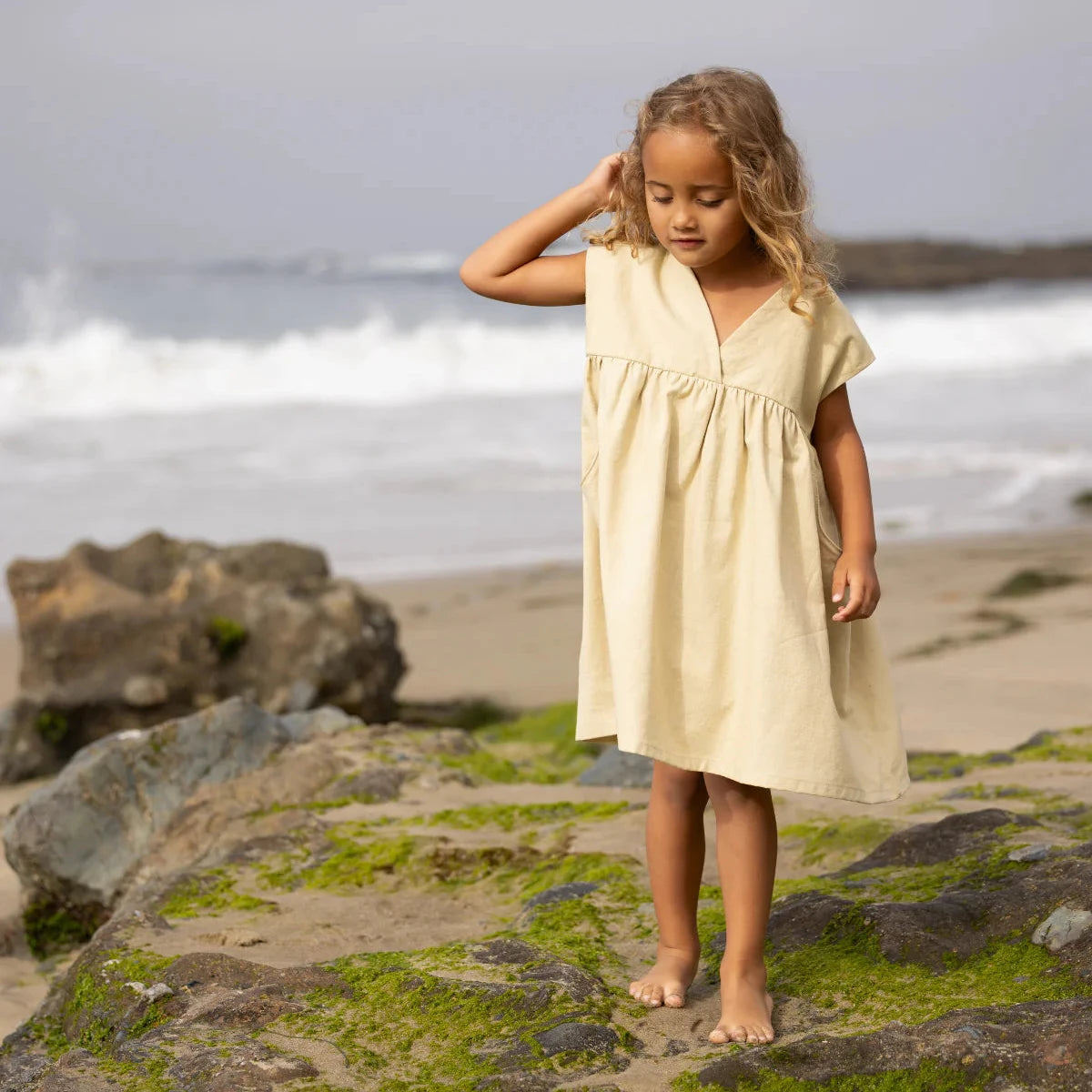 Young girl in a beige dress standing on a rocky beach with ocean waves in the background