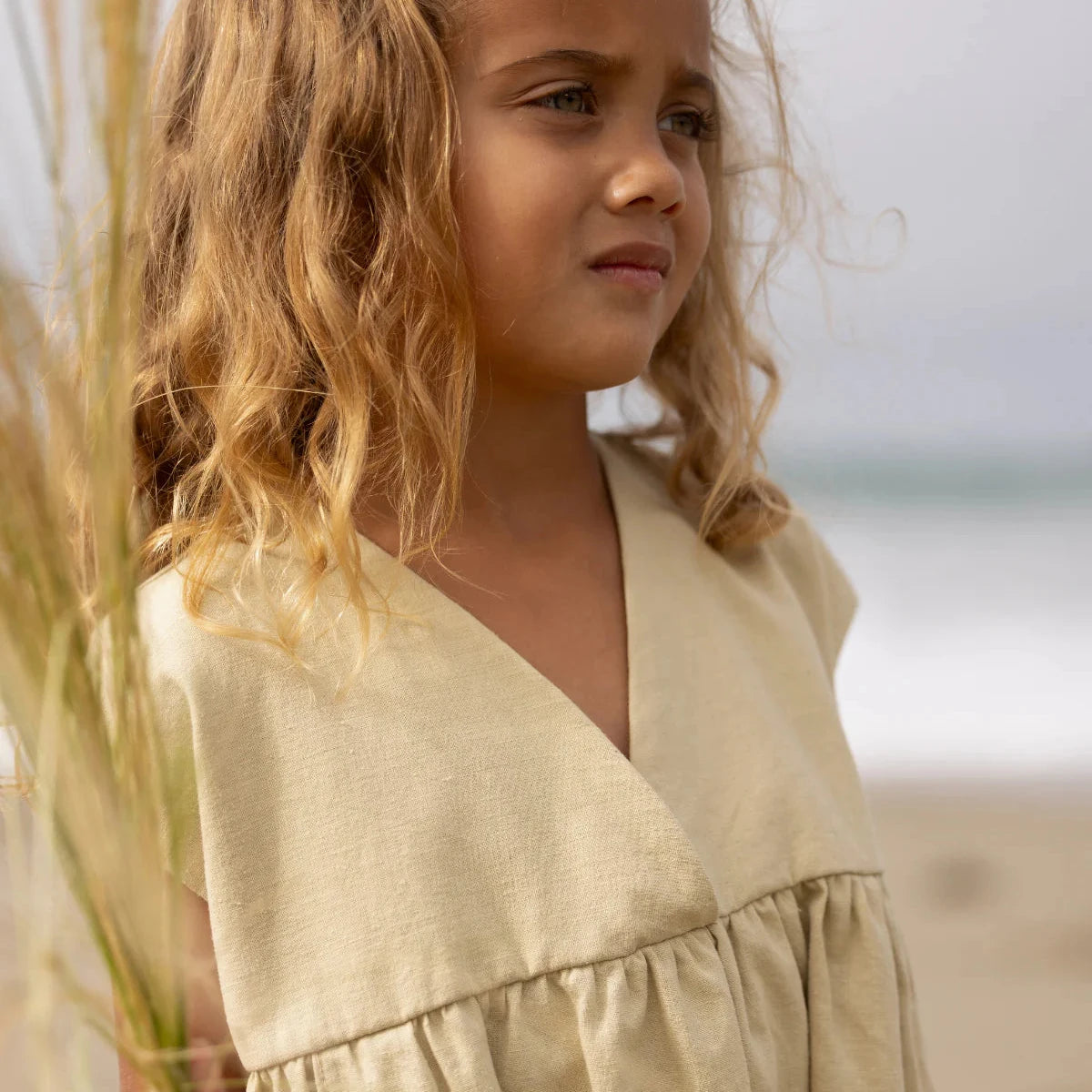 Young girl in a beige dress standing on a rocky beach with ocean waves in the background