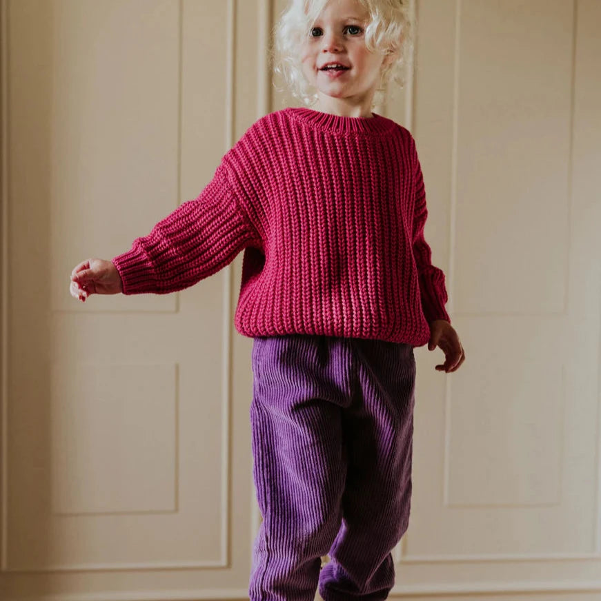 Child wearing a red sweater and purple pants standing on a white platform against a beige paneled wall.