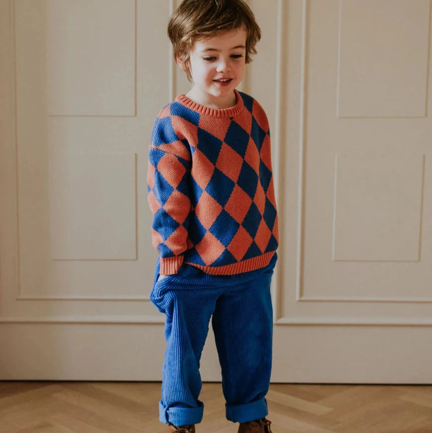 Child wearing a colorful argyle sweater and blue pants standing against a neutral wall.