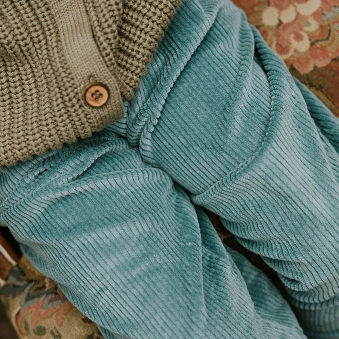 Person wearing a brown knitted cardigan and blue corduroy pants sitting on a patterned rug.