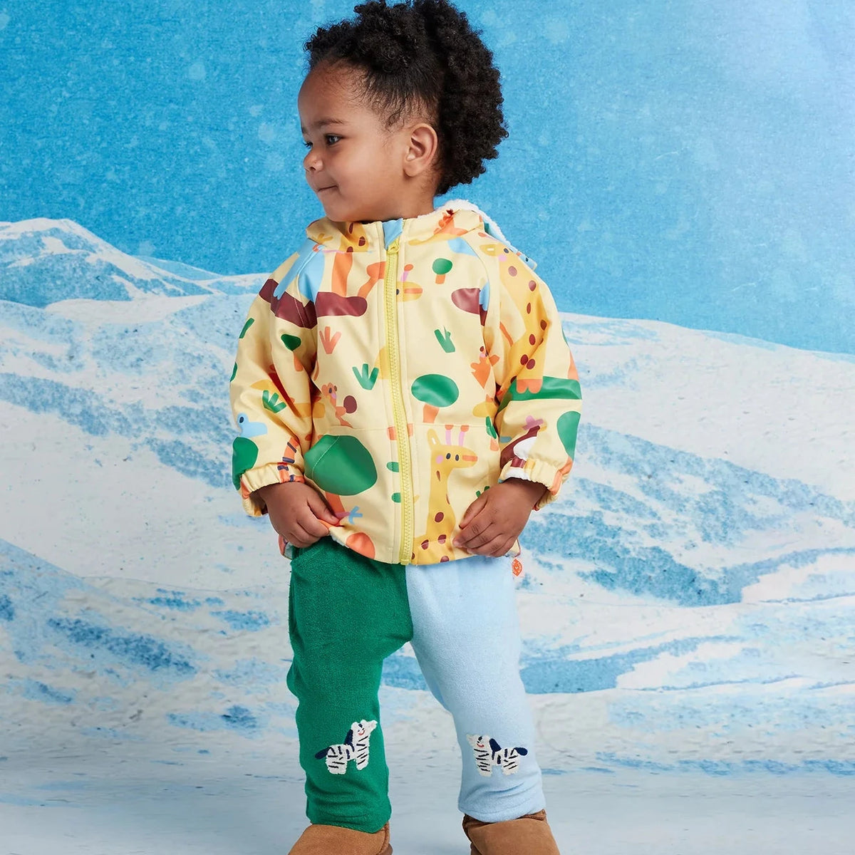 A toddler stands in front of a snowy mountain backdrop wearing a hooded zip-up jacket with a multicolor animal print, green and light blue pants, and brown boots.