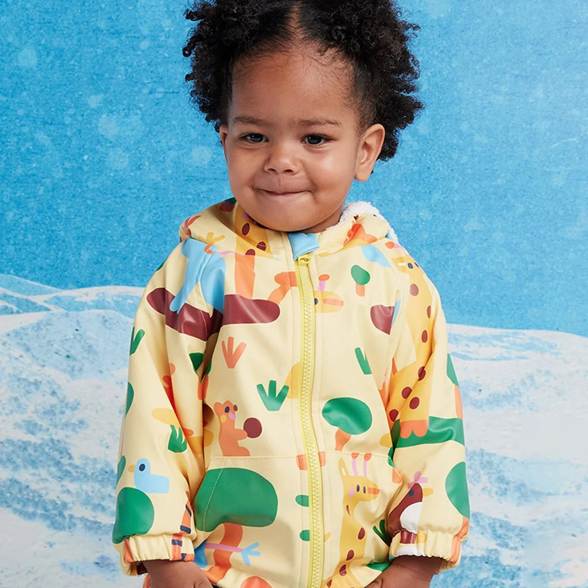A toddler wearing a yellow hooded jacket with a colorful animal and landscape print, standing against a blue and white snowy backdrop.