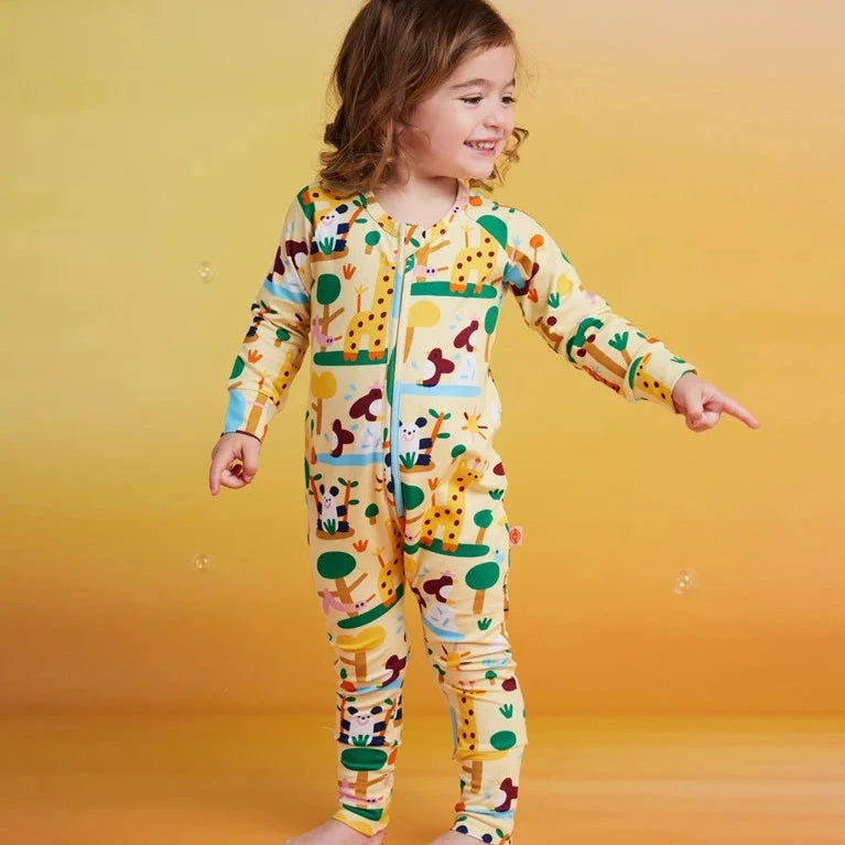A young girl stands barefoot against a warm yellow-to-orange gradient background, wearing a long-sleeve footed zip-up pajama covered in a colorful giraffe and safari animal print.