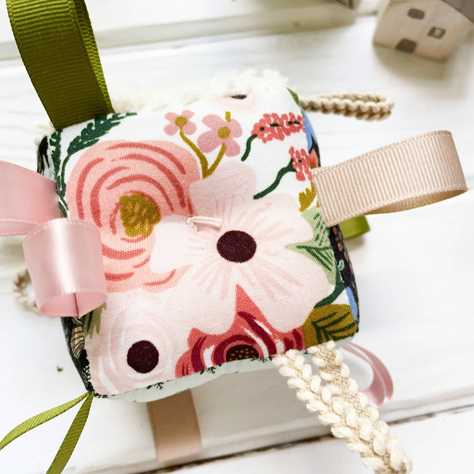 A floral baby rattle block with multiple colors including rose, gold, blush, pale mint, blue, and black, featuring braided loops, ribbons, and a rattle attached to a green ribbon.
