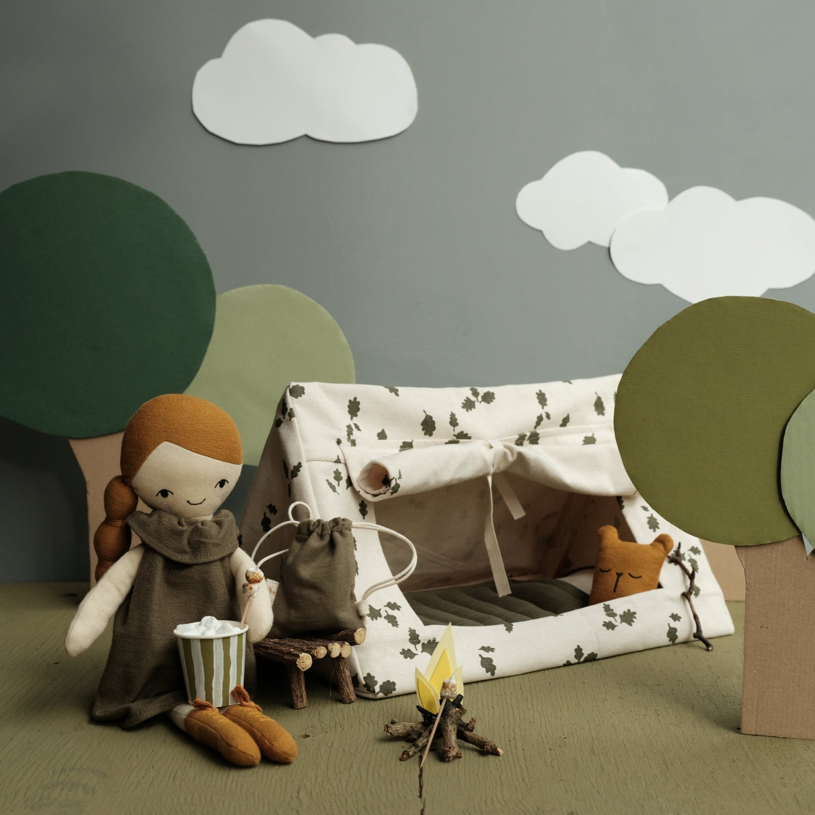 A doll tent with a flashlight, picnic blanket, and a book, set against a background with clouds and trees. A doll is seen sitting beside the tent.