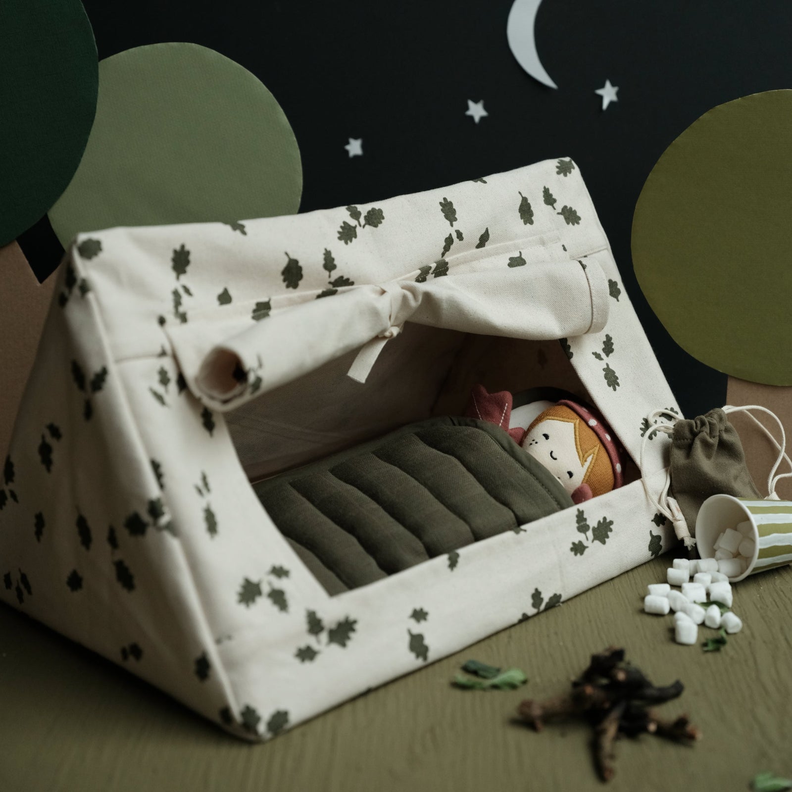 A doll tent with a flashlight, picnic blanket, and a book, set against a background with clouds and trees. A doll is seen sitting beside the tent.